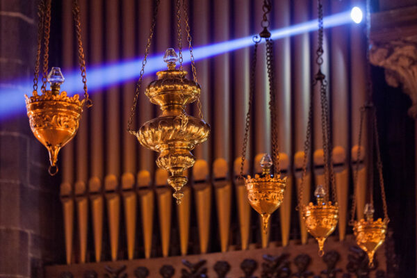Ornate gilded hanging censers suspended by chains in front of cathedral organ pipes, illuminated by warm light and a blue spotlight beam