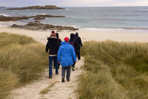 A group of people in winter coats walking through sand dunes towards a rocky beach and sea on a cool, overcast day